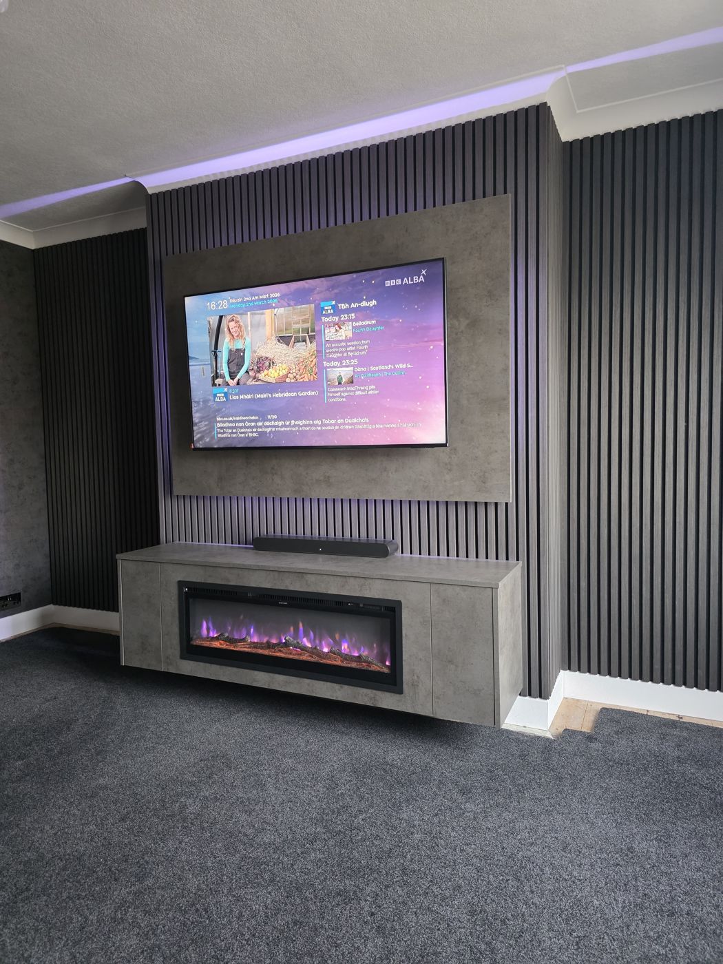 Grey concrete media wall on a black slat wall in Ayrshire, with warm LED lighting, a floating cabinet and an electric fire below the TV, by MPS Media Walls.