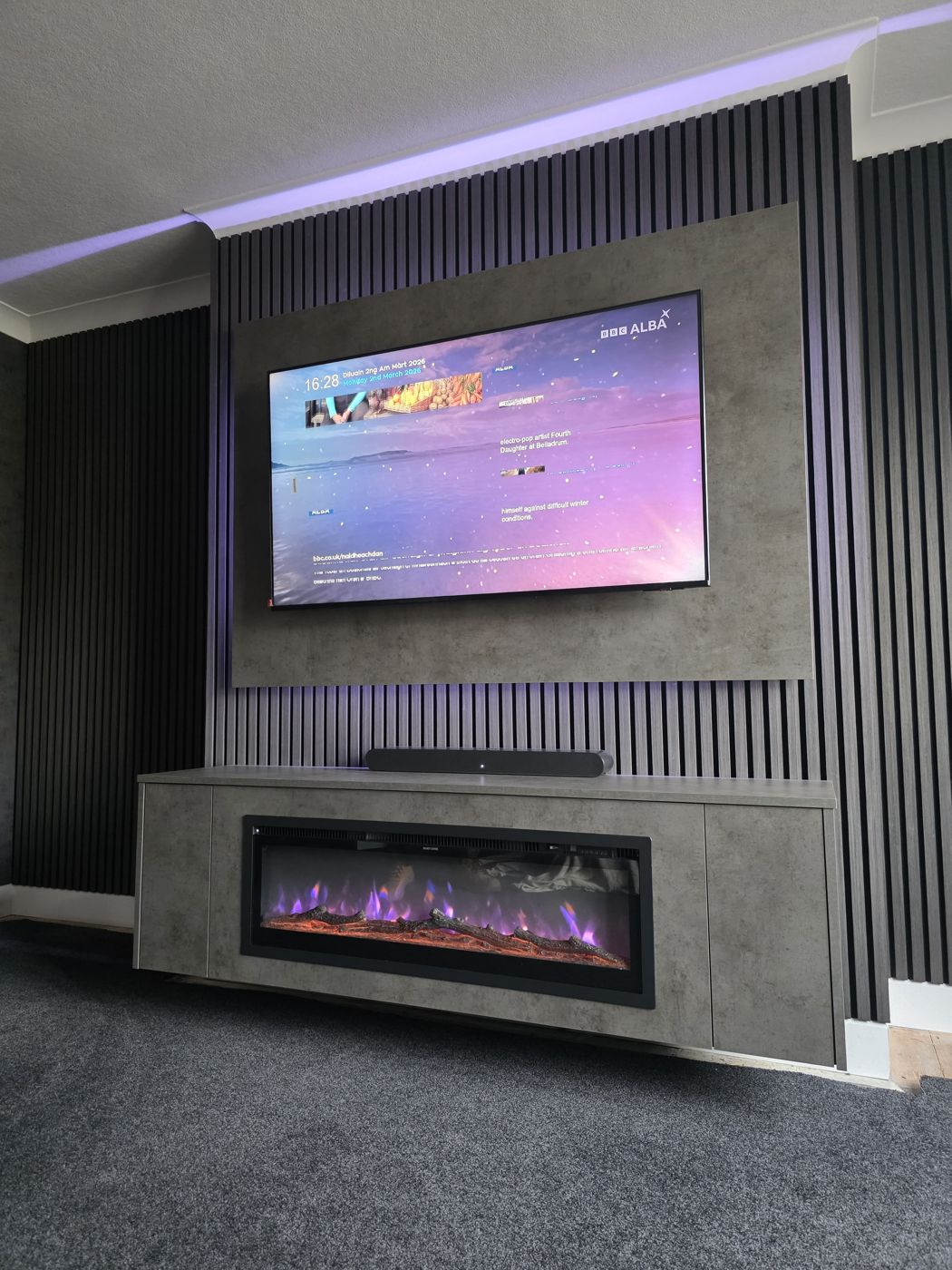 Grey concrete media wall in Ayrshire, angled view, black slats, purple LED glow and electric fire with purple flames.