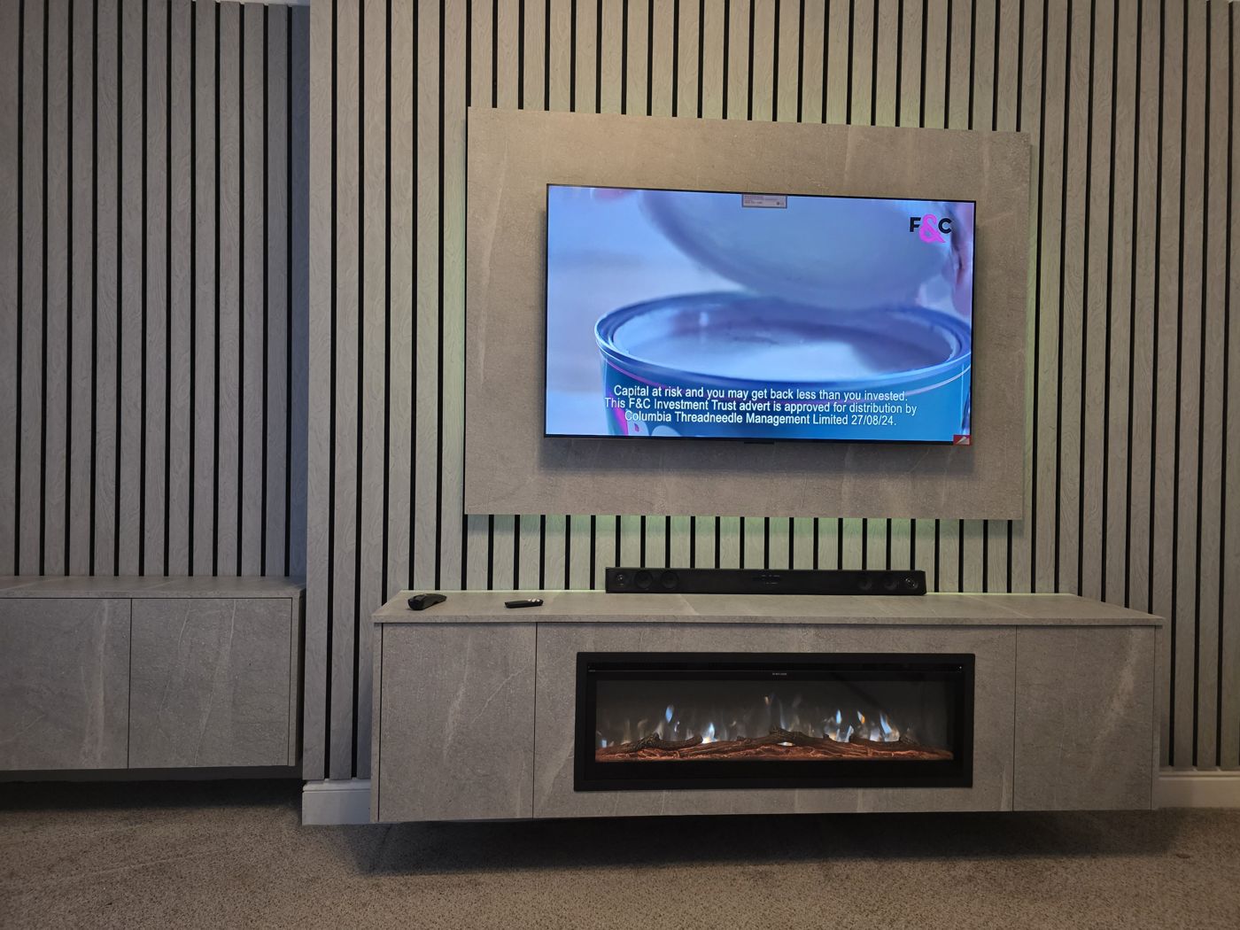 Falkirk media wall, grey stone-effect panel, grey slats, soundbar and long floating cabinet with electric fire below the TV.