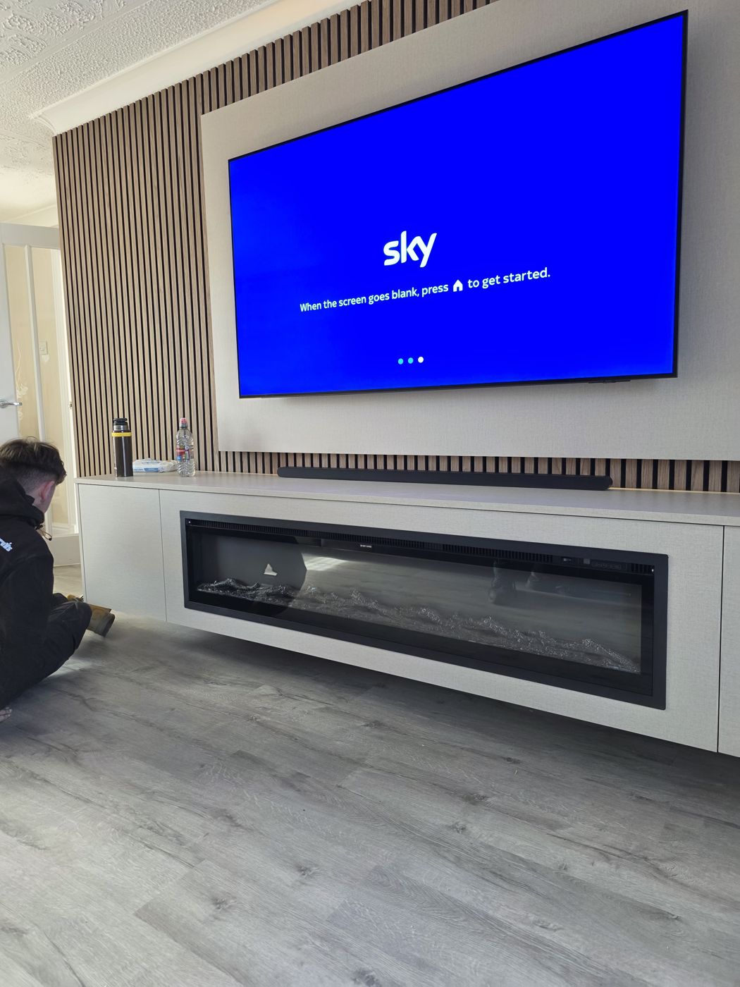 Fife media wall with oak slats, a cream concrete panel, a soundbar and a long floating cabinet with an electric fire below the TV, by MPS Media Walls.
